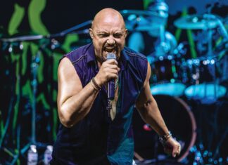 A bald singer in a sleeveless purple shirt sings into a microphone on a blue-lit stage with drums behind him.
