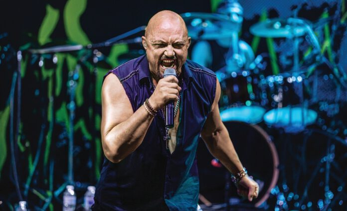 A bald singer in a sleeveless purple shirt sings into a microphone on a blue-lit stage with drums behind him.