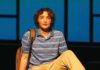 Liar: ‘Dear Evan Hansen’ at Spreckels An actor with curly hair sits on stage wearing a striped polo and khakis, a backpack beside him, against a blue-lit backdrop.