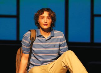 An actor with curly hair sits on stage wearing a striped polo and khakis, a backpack beside him, against a blue-lit backdrop.