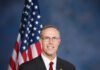 Rep. Jared Huffman smiles in a dark suit and red tie, standing beside an American flag.