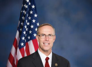 Rep. Jared Huffman smiles in a dark suit and red tie, standing beside an American flag.