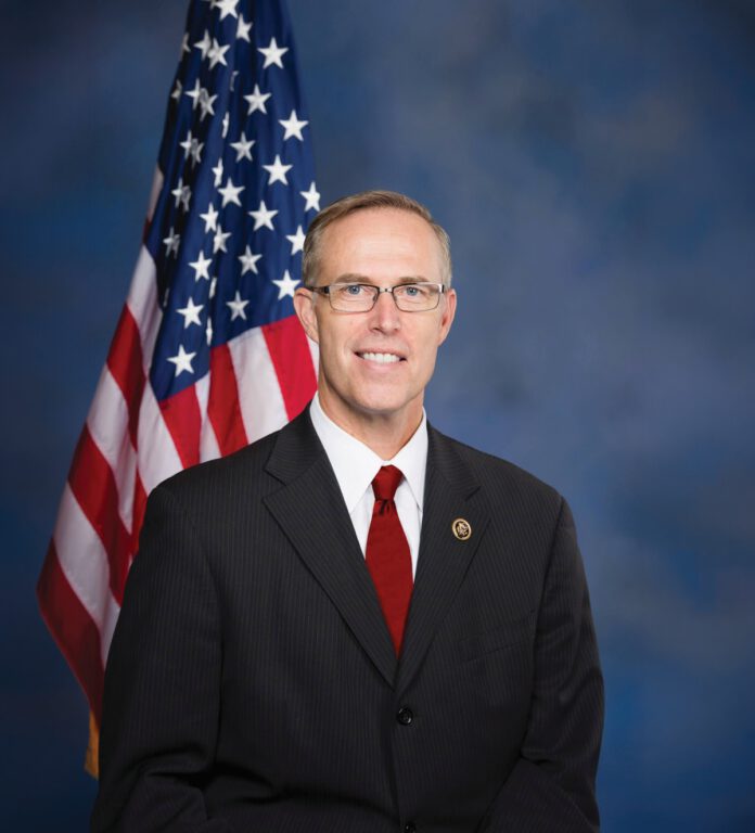 Rep. Jared Huffman smiles in a dark suit and red tie, standing beside an American flag.