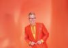 A smiling man with gray hair and black-framed glasses wears an orange blazer, yellow shirt and fancy orange and yellow tie, while standing in front of an orange background.