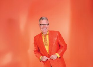 Resort-O-Rama, Altered States and Moving Pictures A smiling man with gray hair and black-framed glasses wears an orange blazer, yellow shirt and fancy orange and yellow tie, while standing in front of an orange background.