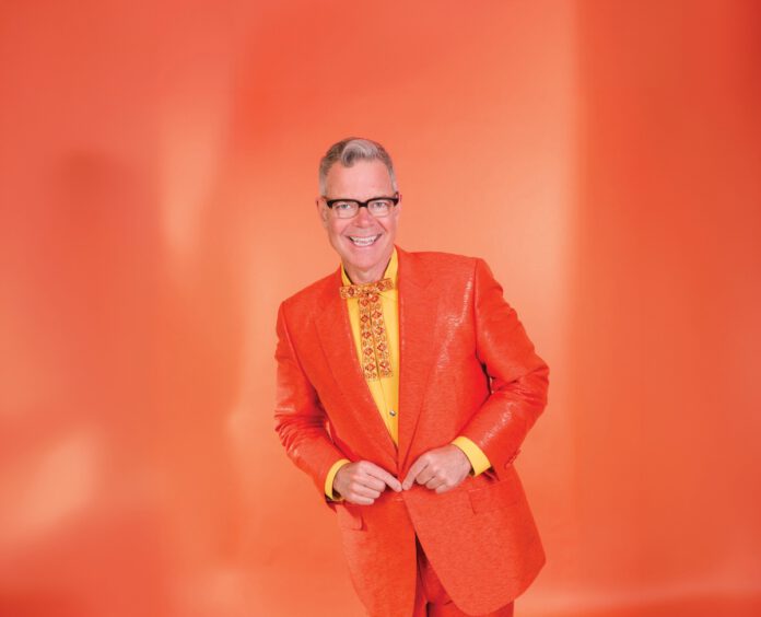 A smiling man with gray hair and black-framed glasses wears an orange blazer, yellow shirt and fancy orange and yellow tie, while standing in front of an orange background.