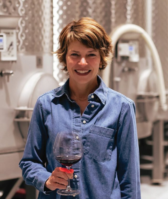 A smiling woman in a denim shirt holds a glass of red wine in front of large stainless steel tanks.
