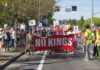People marching on a street carry a red banner reading “NO KINGS” and others behind them hold protest signs.