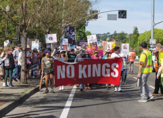 People marching on a street carry a red banner reading “NO KINGS” and others behind them hold protest signs.