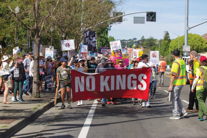 People marching on a street carry a red banner reading “NO KINGS” and others behind them hold protest signs.