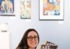 Linked Hands: Penelope Lenaerts of Art Trails Open Studios A smiling woman with long brown hair, eyeglasses and a nose ring sits on a wooden bench, holding two catalogs titled “Art at the Source: Open Studios.”