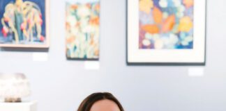 A smiling woman with long brown hair, eyeglasses and a nose ring sits on a wooden bench, holding two catalogs titled “Art at the Source: Open Studios.”
