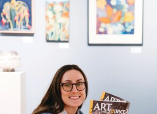 A smiling woman with long brown hair, eyeglasses and a nose ring sits on a wooden bench, holding two catalogs titled “Art at the Source: Open Studios.”