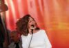Divine Divas, Spanglish and Haunted Wine A woman with curly auburn hair wears a white dress and sings into a microphone, keeping her eyes closed.