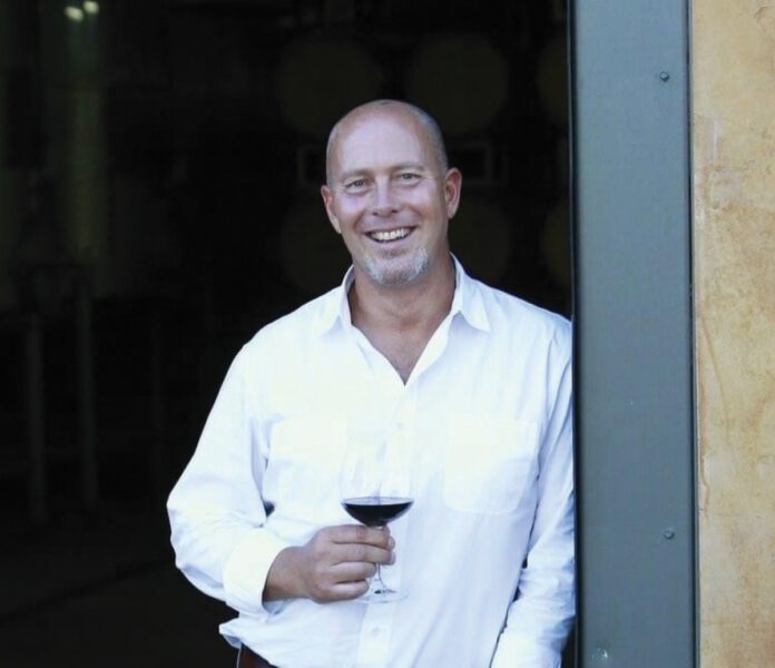 A smiling man dressed in a white shirt holds a glass of red wine as he leans against an open door.