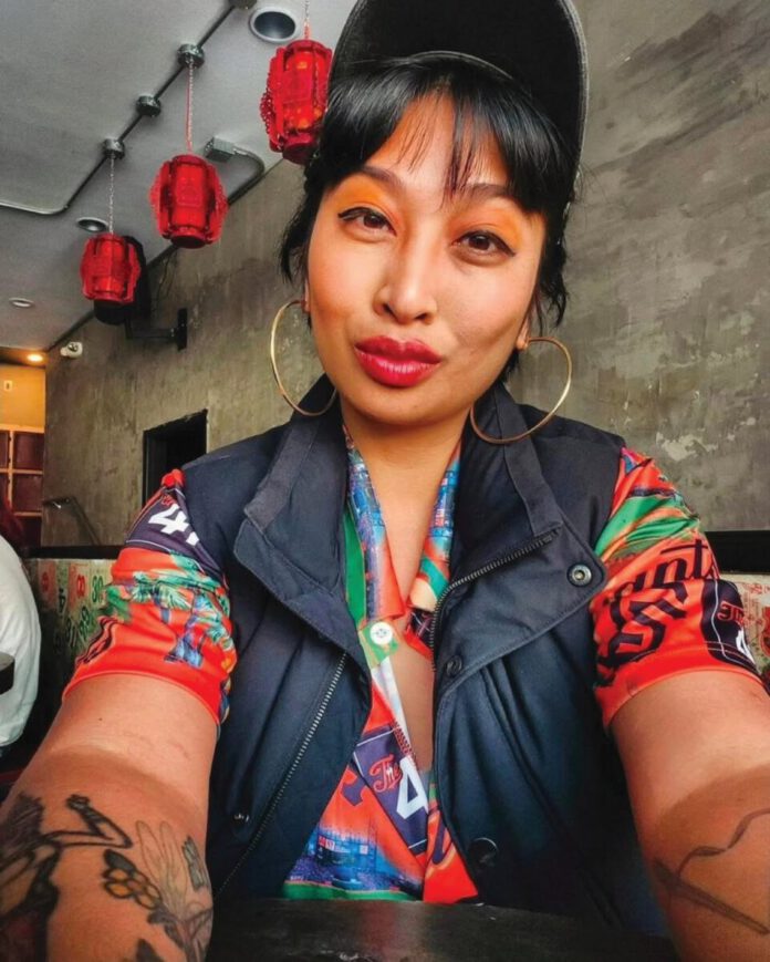 A woman with black bangs is wearing a black cap, an orange shirt, a black vest and gold hoop earrings and she sports orange eyeshadow, red lipstick and arm tattoos.