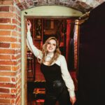 Sound Decisions: Announcing Our 2026 North Bay Music Awards Winners A smiling woman with long blonde hair, a white shirt and black leather pants leans against a brick wall.
