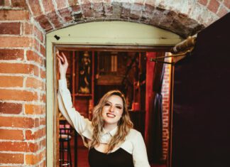 A smiling woman with long blonde hair, a white shirt and black leather pants leans against a brick wall.