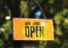 A yellow sign reads “we are OPEN,” and hangs on twine in front of a window.