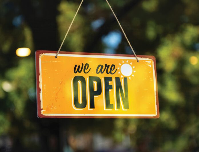 A yellow sign reads “we are OPEN,” and hangs on twine in front of a window.