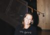 A smiling woman with shoulder-length hair wearing a black shirt that reads “une femme” stands in front of a wood staircase.