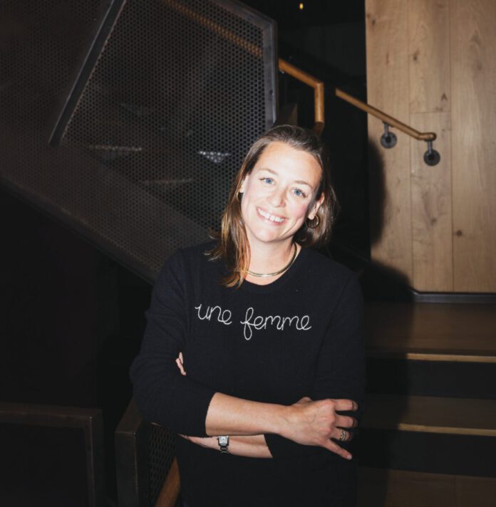 A smiling woman with shoulder-length hair wearing a black shirt that reads “une femme” stands in front of a wood staircase.