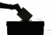 In silhouette, a hand drops a folded ballot into a black box.
