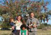 A smiling woman holding a toddler, a girl holding a bottle and a smiling man holding bottles stand in front of an apple tree full of red apples.