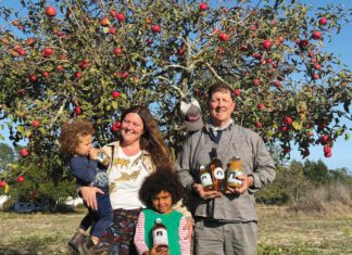 Family Tree: Paul Kolling of Nana Mae’s Organics A smiling woman holding a toddler, a girl holding a bottle and a smiling man holding bottles stand in front of an apple tree full of red apples.