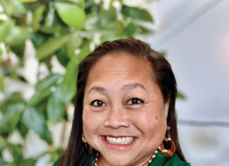 A smiling woman with long, straight dark hair is wearing gold jewelry and a green blouse.