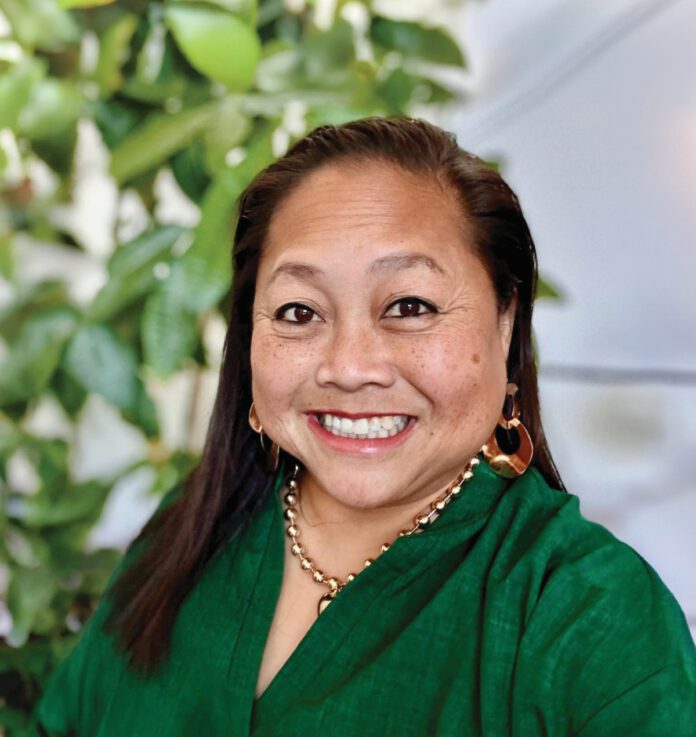 A smiling woman with long, straight dark hair is wearing gold jewelry and a green blouse.