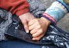 Two hands from two different people are clasped over a black plastic bag. One person has a wrinkled hand, while the other hand is smooth and younger-looking.