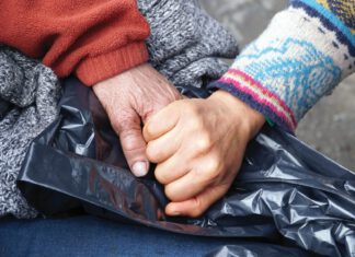 Two hands from two different people are clasped over a black plastic bag. One person has a wrinkled hand, while the other hand is smooth and younger-looking.