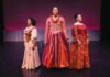 Three women in Elizabethan-style gowns stand in a row on a dark stage in front of a magenta background.