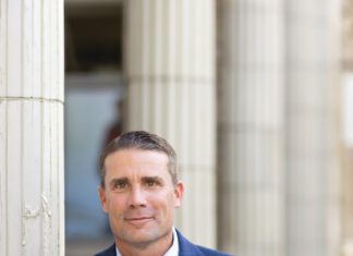 A smiling man with short brown hair wearing a navy plaid blazer and light blue shirt leans against a white column.
