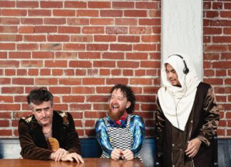 Three men pose in front of a red brick wall. One wears a brown jacket and leans on a wooden table. Another with curly hair and a beard wears checkered overalls and is laughing. The third stands, wearing a white hooded scarf and using headphones.