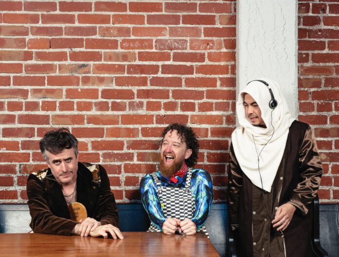 Three men pose in front of a red brick wall. One wears a brown jacket and leans on a wooden table. Another with curly hair and a beard wears checkered overalls and is laughing. The third stands, wearing a white hooded scarf and using headphones.
