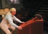 On stage, an older man in a cream vest and striped shirt bends over and grabs a red wood box that has a man's head poking out of a hole.