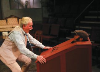 All Talk: G.B. Shaw’s ‘Misalliance’ at 6th Street On stage, an older man in a cream vest and striped shirt bends over and grabs a red wood box that has a man's head poking out of a hole.