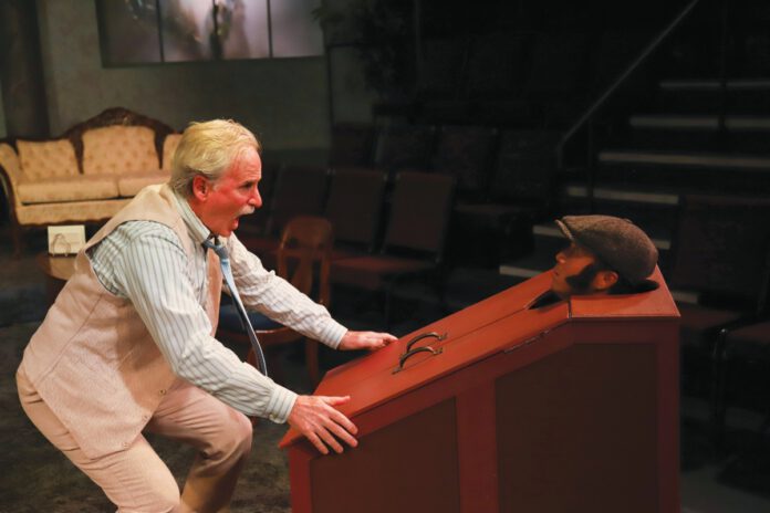 On stage, an older man in a cream vest and striped shirt bends over and grabs a red wood box that has a man's head poking out of a hole.