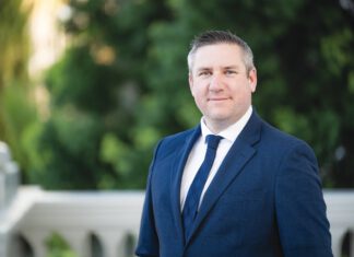A man with short brown hair and graying temples wearing a blue plaid suit and a navy tie stands outdoors in front of a white balustrade and green foliage.