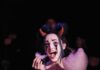 A woman in clown makeup wearing red devil horns and a spiderweb collar pulls on pink cotton candy in a dark room.