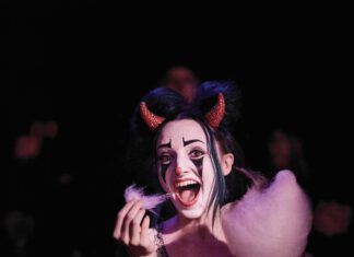 A woman in clown makeup wearing red devil horns and a spiderweb collar pulls on pink cotton candy in a dark room.