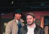 A man with a beard wears a cap and leather jacket and has a cigarette in his mouth while standing in front of a man in a fedora and trench coat. Both are on a stage set.