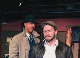 Suspicious Minds: ‘Night Must Fall’ in Yountville A man with a beard wears a cap and leather jacket and has a cigarette in his mouth while standing in front of a man in a fedora and trench coat. Both are on a stage set.