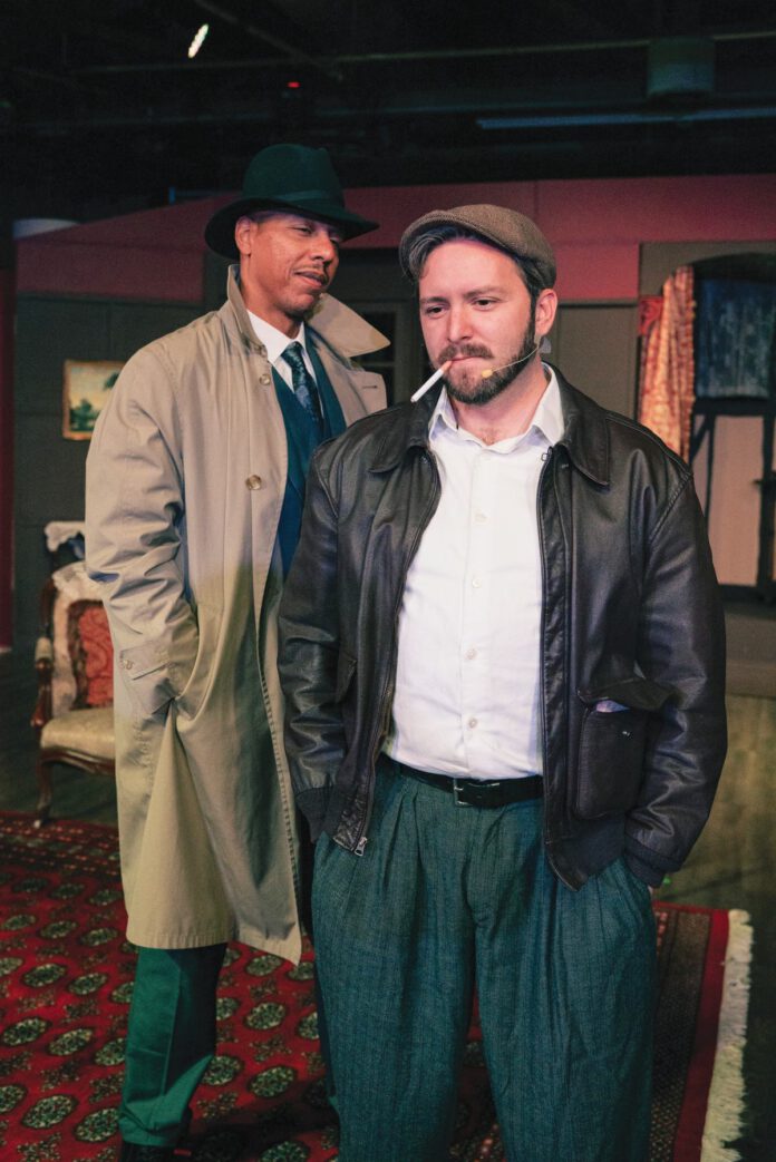A man with a beard wears a cap and leather jacket and has a cigarette in his mouth while standing in front of a man in a fedora and trench coat. Both are on a stage set.