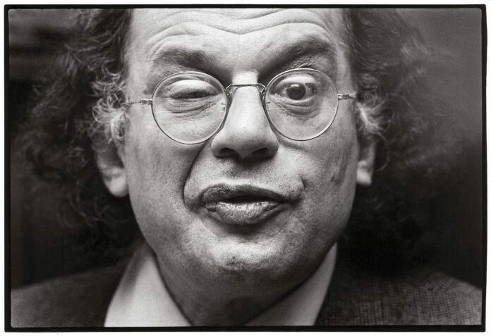 A black-and-white close-up of a man with curly hair and round wire-rimmed glasses, who has one eye squinted and his lips pursed.