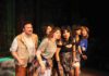 Six performers in ’80s-style costumes, a man and five women, stand and look forward on the stage.