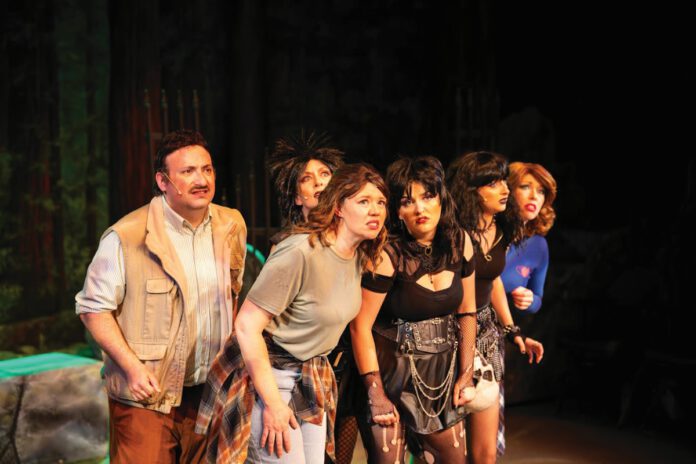 Six performers in ’80s-style costumes, a man and five women, stand and look forward on the stage.